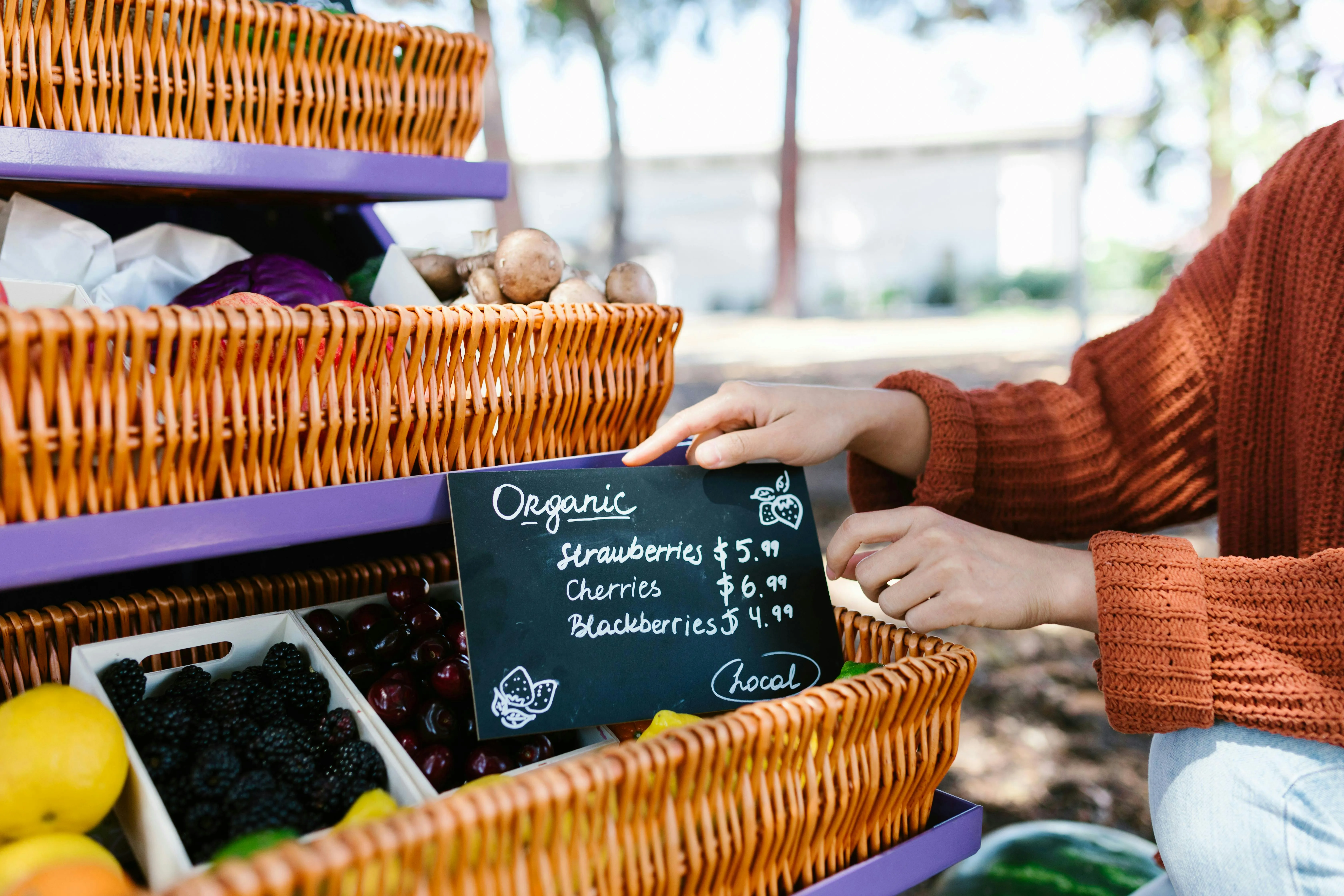 Person arranging a handwritten chalkboard sign with organic produce prices on a wicker display stand, showcasing fresh fruits like strawberries, cherries, and blackberries at an outdoor market.